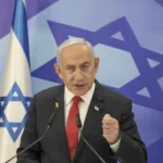 Portrait of Benjamin Netanyahu in a dark suit and blue tie, with an Israeli flag blurred in the background.