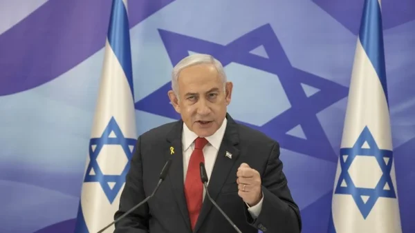 Portrait of Benjamin Netanyahu in a dark suit and blue tie, with an Israeli flag blurred in the background.