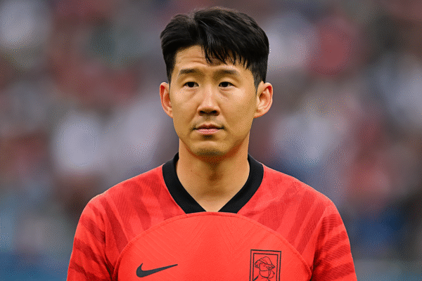 Son Heung-min wearing a red South Korean football jersey, standing on the field with a focused expression and a blurred stadium background.